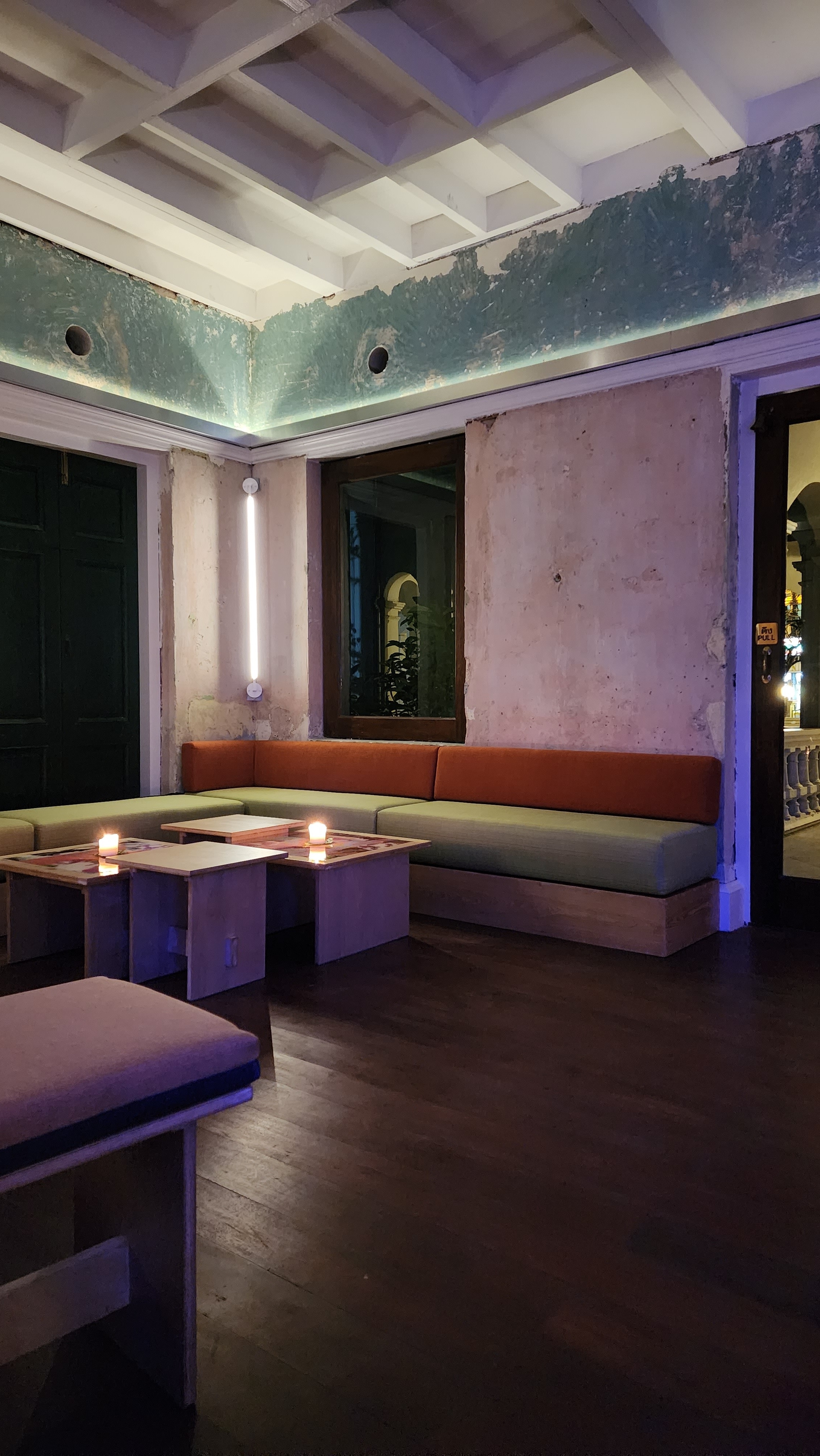 The warm interior of a cafe. The walls are pink, the booths lining the outer walls green and red. Tables in the corner of the booths hold burning candles.